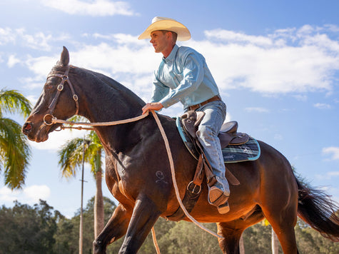 Cowboy riding brown horse wearing Ariat clothing and boots with blue sky and palm trees background.