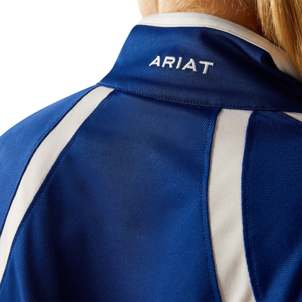 Woman wearing a blue and white long-sleeve athletic jacket with Ariat branding with a white cap on a white background