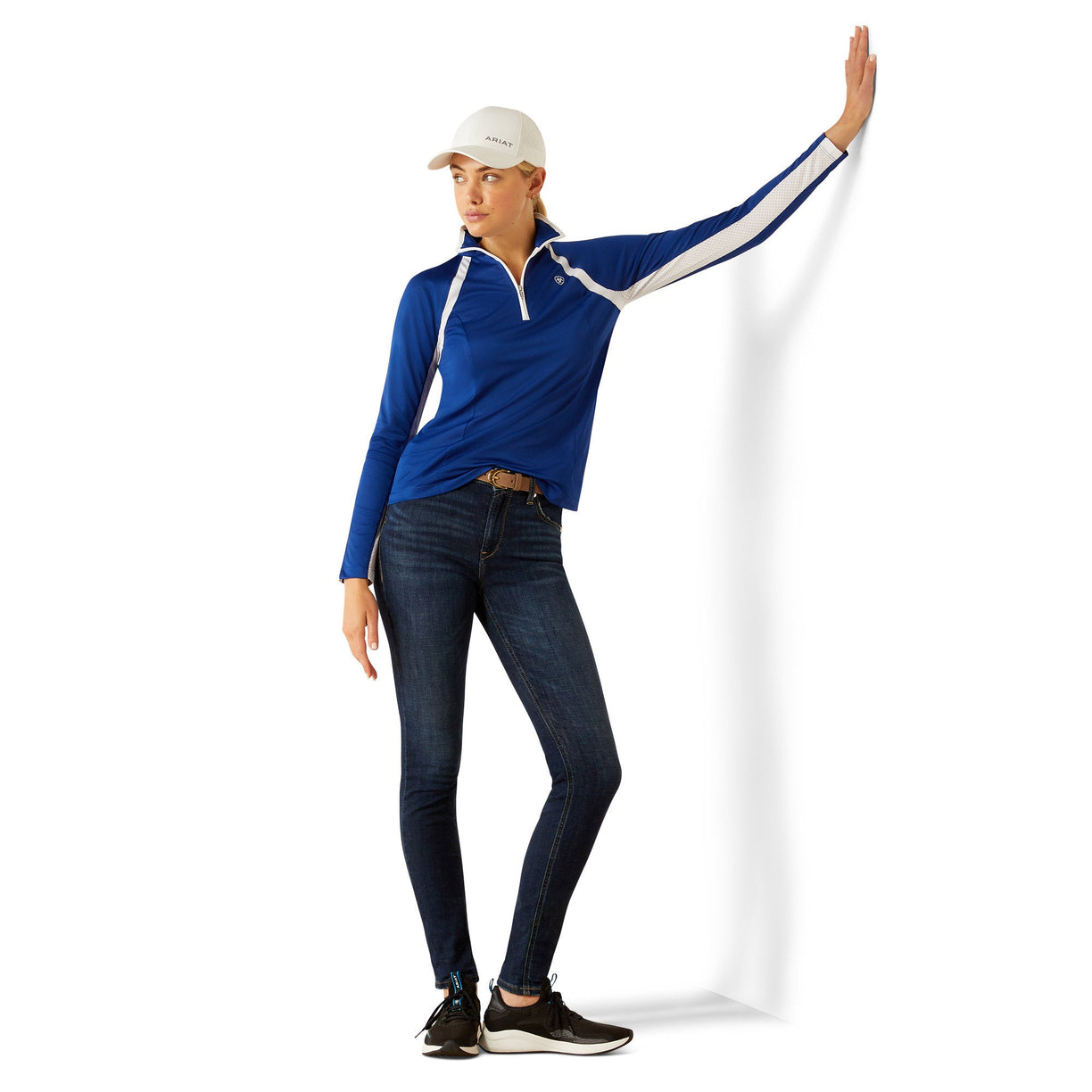 Woman wearing a blue and white long-sleeve athletic jacket with Ariat branding with a white cap on a white background