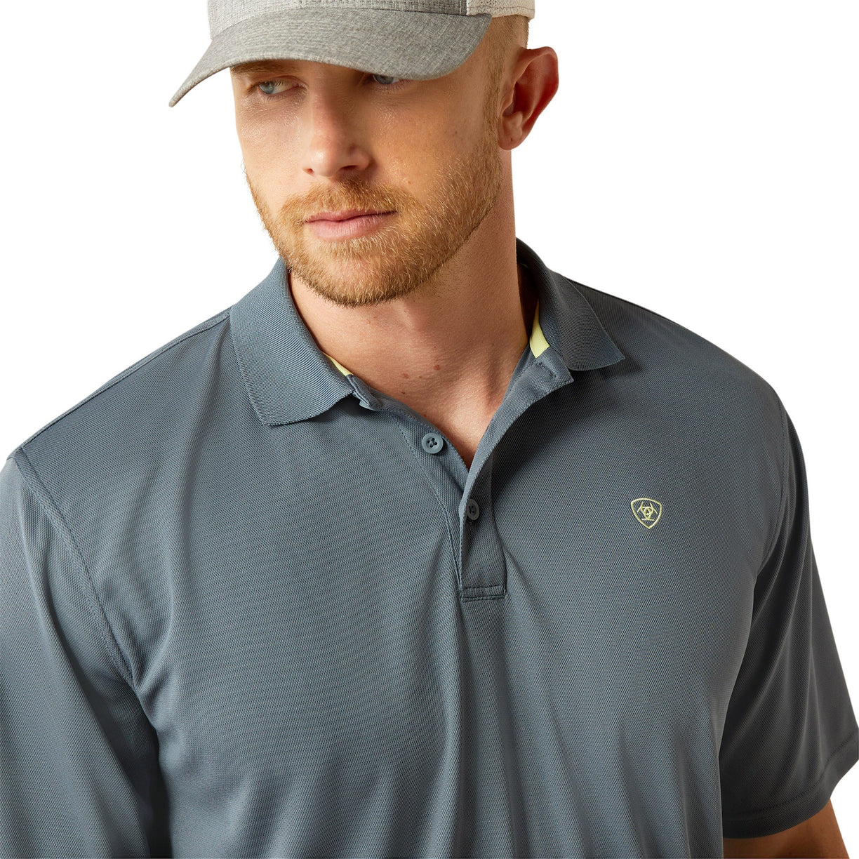 Man wearing a blue gray polo shirt with Ariat branding and cap on a white background
