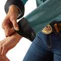 Person wearing a patterned shirt and blue jeans, adjusting a black device on their wrist against a white background.