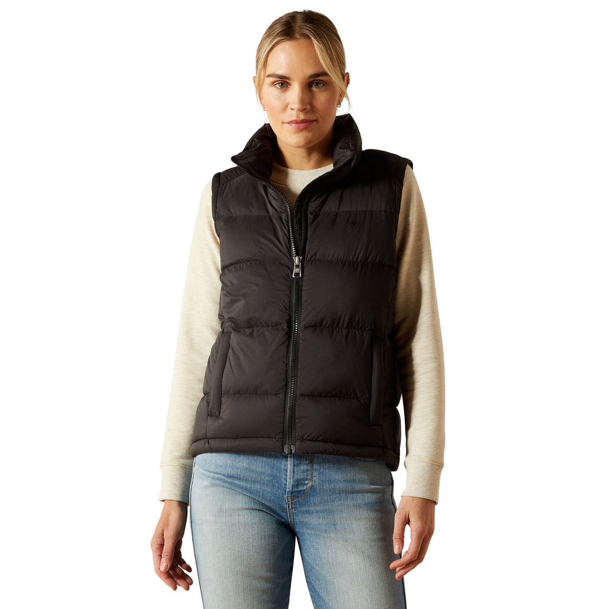 Woman wearing a black puffer vest over a beige sweater with blue jeans on a white background
