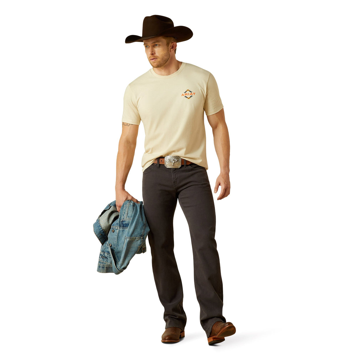 Man wearing a beige t-shirt, dark jeans, and a cowboy hat holding a denim jacket on a white background
