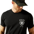 Man wearing a black t-shirt with Ariat logo and a camouflage cap on a white background