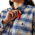 Person wearing a blue plaid shirt with a visible Ariat logo, holding a red pen.