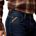 Person wearing blue jeans with Ariat logo, brown plaid shirt on a white background