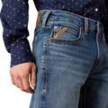 Close-up of blue jeans with a brand label, worn by a person wearing a patterned shirt.