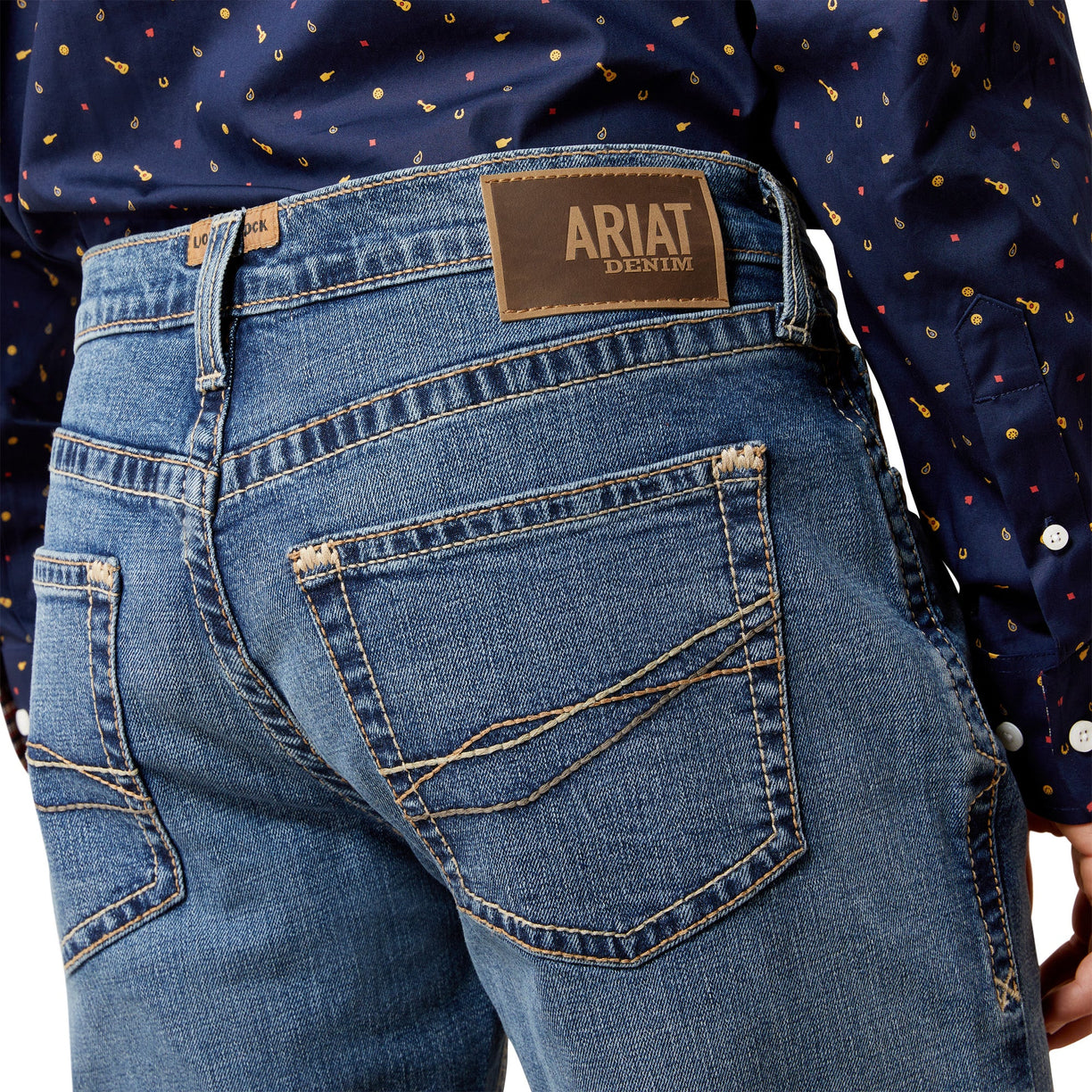 Close-up of blue jeans with Ariat denim label, worn by a person in a patterned shirt.