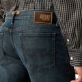 Person wearing blue jeans with Ariat denim label, plaid shirt visible