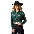 Woman wearing a green cowboy shirt with white stitching, black cowboy hat, and dark jeans on a beige background.
