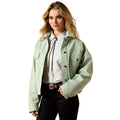A woman modeling a light sage green cropped utility jacket over a white collared shirt with a tie, paired with black pants and a brown belt, on a solid white background.