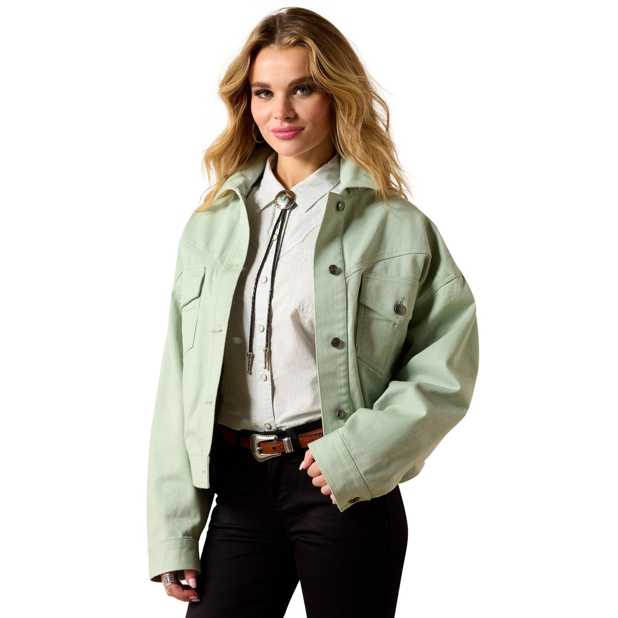 A woman modeling a light sage green cropped utility jacket over a white collared shirt with a tie, paired with black pants and a brown belt, on a solid white background.