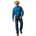 A man wearing a bright blue long-sleeve western shirt, dark blue jeans, a brown belt, and a beige cowboy hat, standing against a plain white background.