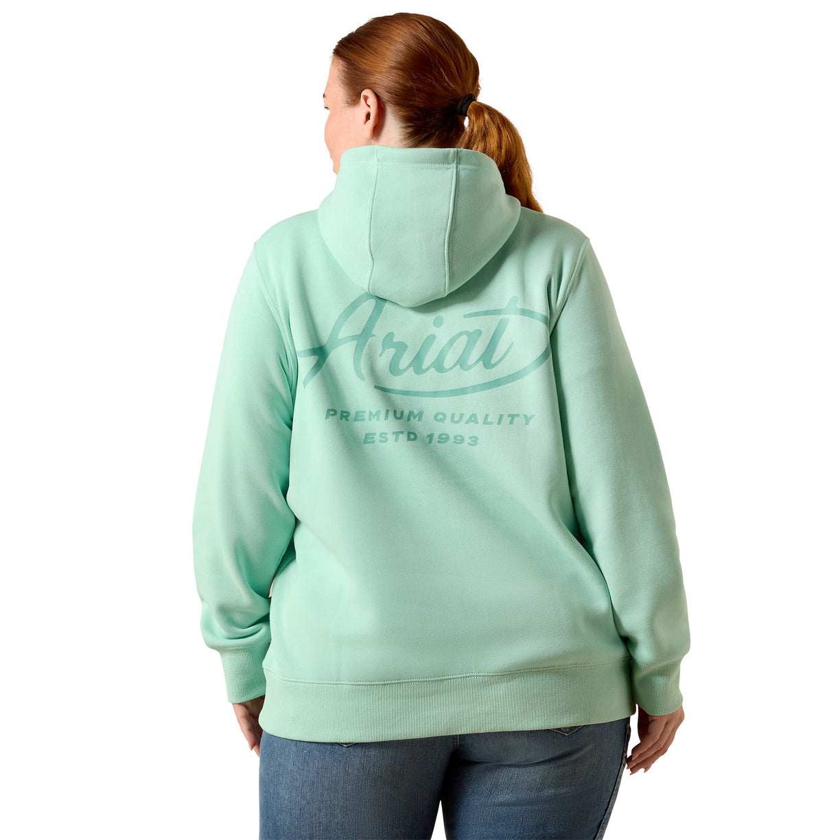 A woman viewed from the back wearing a mint green hoodie with a large 'Ariat' logo printed on the back, paired with blue jeans against a plain white background.