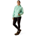 A woman modeling a light mint green hooded sweatshirt with black leggings and brown boots, standing against a plain white studio background.