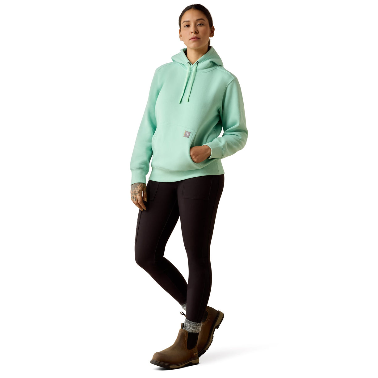 A woman modeling a light mint green hooded sweatshirt with black leggings and brown boots, standing against a plain white studio background.