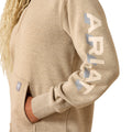 Close-up of a person wearing a heather tan pullover hoodie with a large vertical ARIAT logo printed on the sleeve in cream and light blue, plus a small brand patch on the pocket.