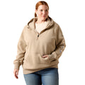 A woman modeling a beige half-zip hoodie with a drawstring hood and kangaroo pocket, paired with blue jeans, against a plain white background.
