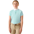 A young girl wearing a light blue equestrian polo shirt with a white collar, paired with beige riding pants and a patterned belt