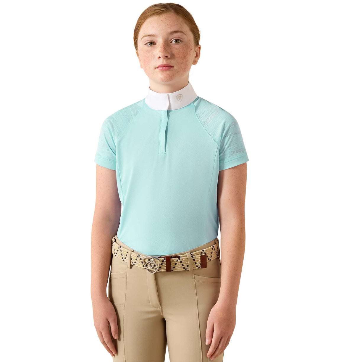A young girl wearing a light blue equestrian polo shirt with a white collar, paired with beige riding pants and a patterned belt