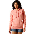 A woman modeling a coral pink pullover hoodie with a small red shield logo on the left chest, paired with denim shorts, against a plain white background.