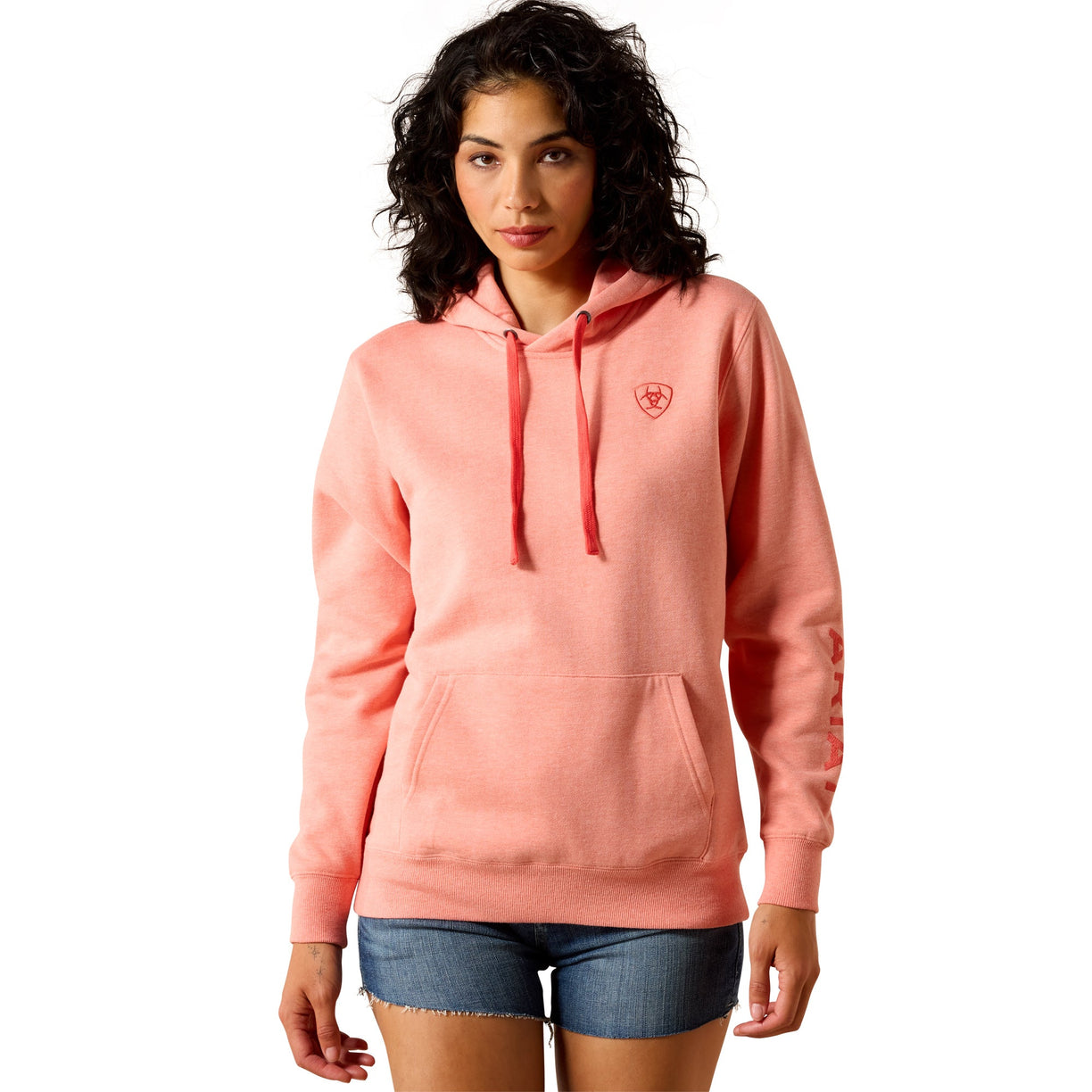 A woman modeling a coral pink pullover hoodie with a small red shield logo on the left chest, paired with denim shorts, against a plain white background.