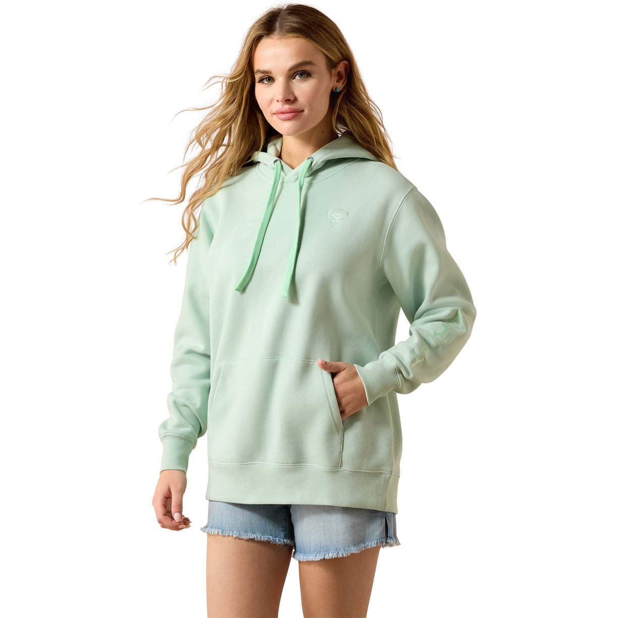 A woman modeling a light mint green pullover hoodie with matching drawstrings and a small tonal chest logo, paired with denim shorts against a plain white background.