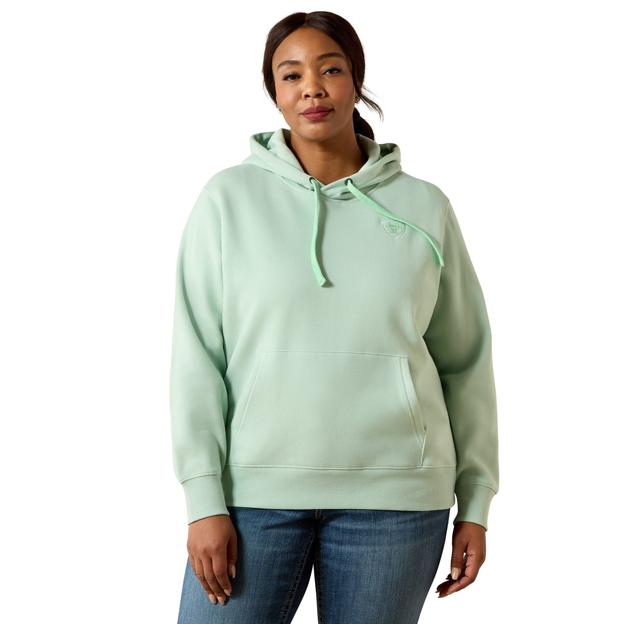 A woman modeling a sage green pullover hoodie with matching drawstrings, paired with blue jeans, against a plain white background.