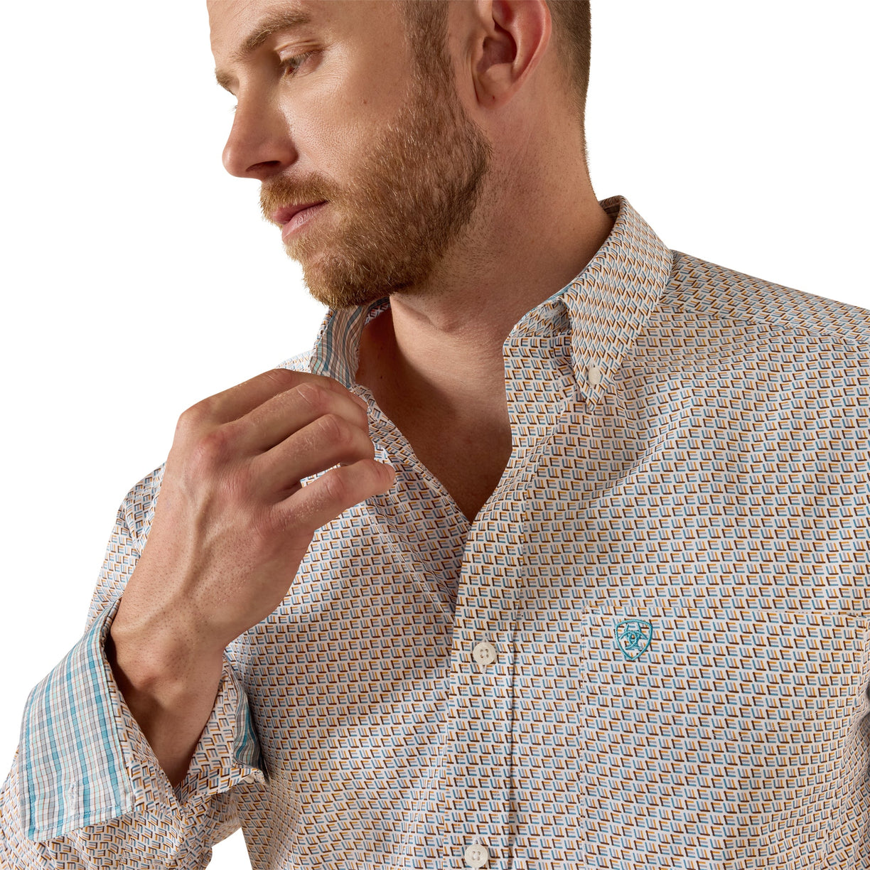 A close-up of a man wearing a light beige button-down shirt with a small blue geometric pattern, adjusting the collar against a white background.