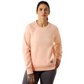 A woman modeling a light peach crewneck sweatshirt with raglan sleeves and a small black brand patch near the lower hem, paired with dark gray leggings against a plain white background.