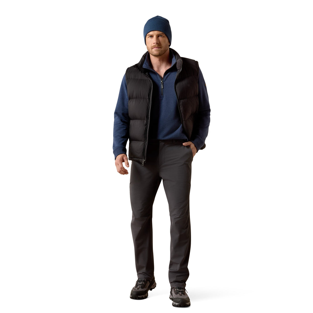 Man wearing a blue beanie, black vest, blue shirt, gray pants, and black shoes on a white background