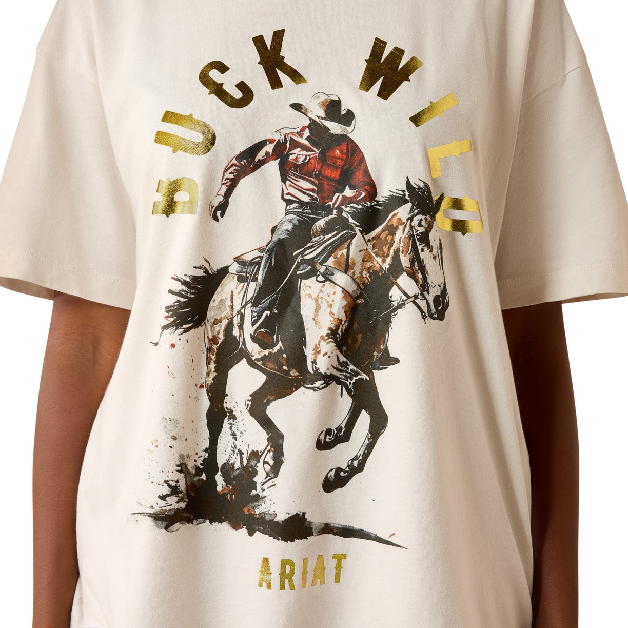 A close-up of a cream-colored t-shirt featuring a graphic print of a cowboy riding a bucking horse, with 'BUCK WILD' written in distressed gold letters and 'ARIAT' at the bottom.