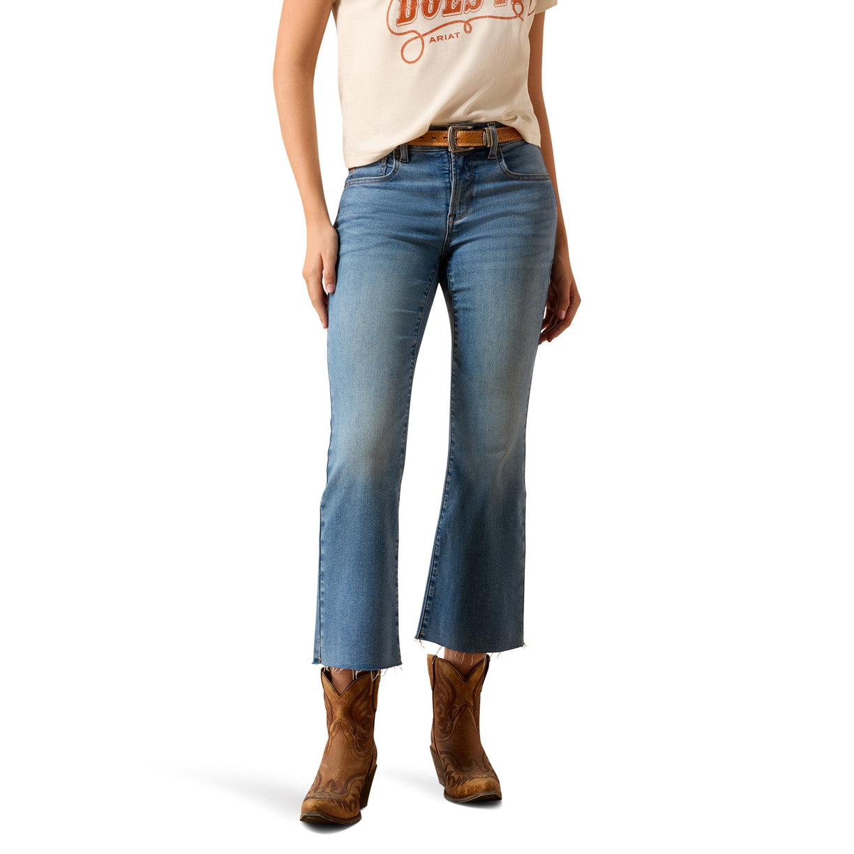 A model wearing a pair of medium-wash blue cropped flare jeans with a brown leather belt, paired with a cream graphic t-shirt and brown cowboy boots, shown against a plain white background.