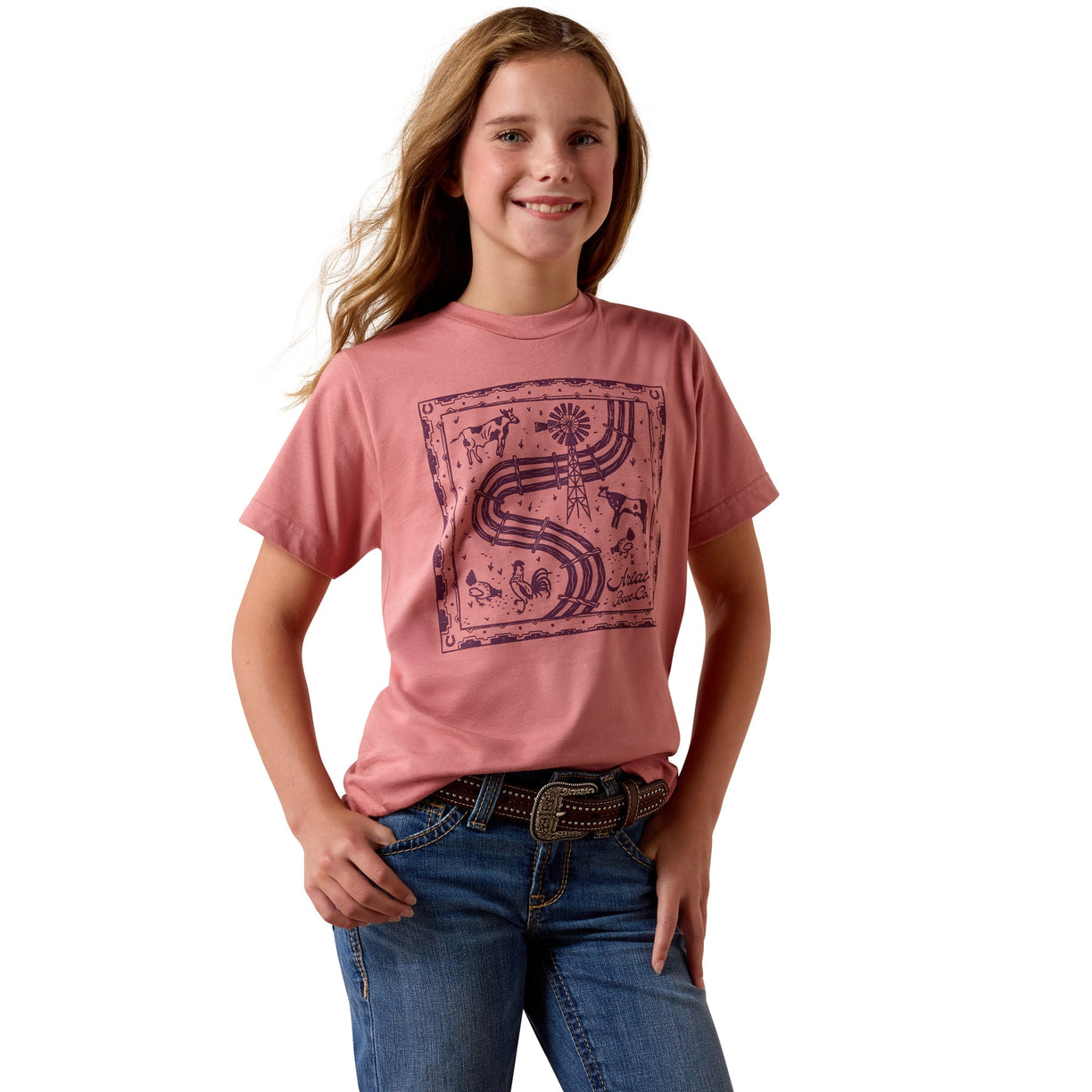 A young girl with long light brown hair smiling, wearing a dusty pink short-sleeve t-shirt with a dark purple graphic print on the front, paired with blue jeans