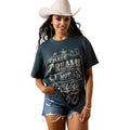 A woman wearing a dark teal graphic t-shirt with distressed denim shorts and a white cowboy hat. The t-shirt features a western-themed graphic with the text 'CHASE DREAMS NOT Cowboys'.