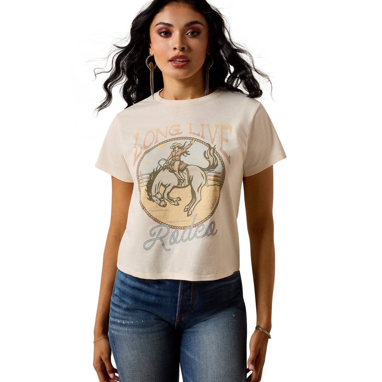Model wearing a beige Ariat t-shirt with a graphic design and text, standing 