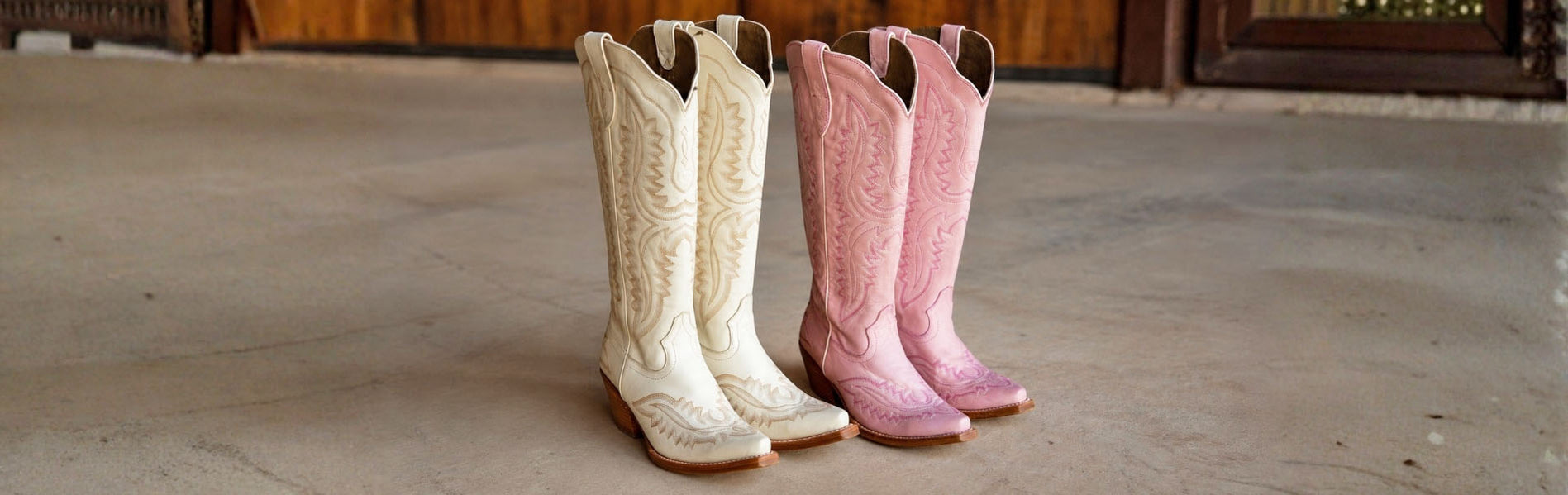 Two pairs of tall western Casanova cowgirl boots standing side by side on a concrete floor. In white and pink with western details.