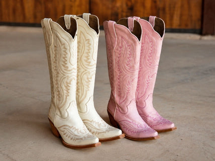 Two pairs of tall western Casanova cowgirl boots standing side by side on a concrete floor. In white and pink with western details.