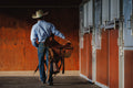 Cowboy walking with a saddle in a stable