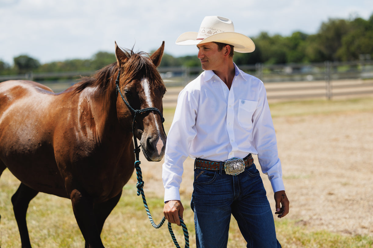 Man in cowboy hat and white shirt standing next to a brown horse in an outdoor setting