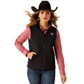 Woman wearing a black vest over a pink shirt with a logo, blue jeans, and a black cowboy hat on a white background.