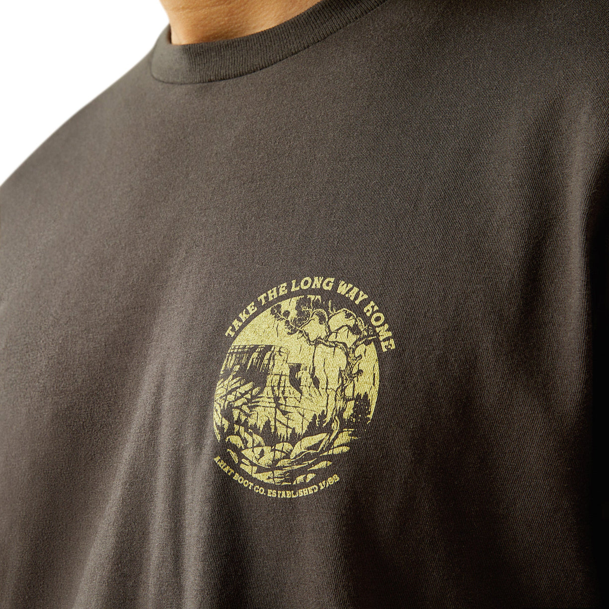 Man wearing a brown t-shirt with a scenic graphic on the back, standing against a white background