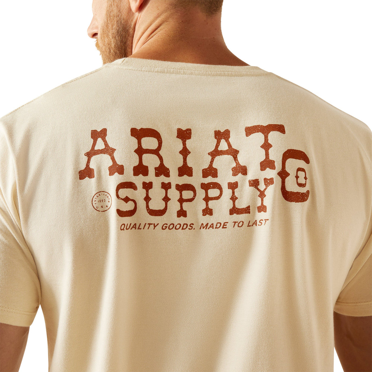 Man wearing a beige t-shirt with 'Ariat Supply' logo on the back, standing against a white background.