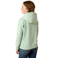 A woman with red hair is shown from the back wearing a light sage green pullover hoodie and blue jeans against a plain white background.