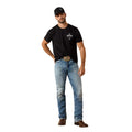 Man wearing a black t-shirt, blue jeans, and a cap against a beige wall.