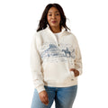 A woman modeling a cream-colored quarter-zip hoodie with a blue desert landscape graphic on the front, paired with blue jeans, against a plain white background.