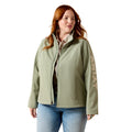 A woman modeling a sage green zip-up jacket with vertical 'AERAT' branding on the sleeve, paired with a patterned shirt and blue jeans against a white background.