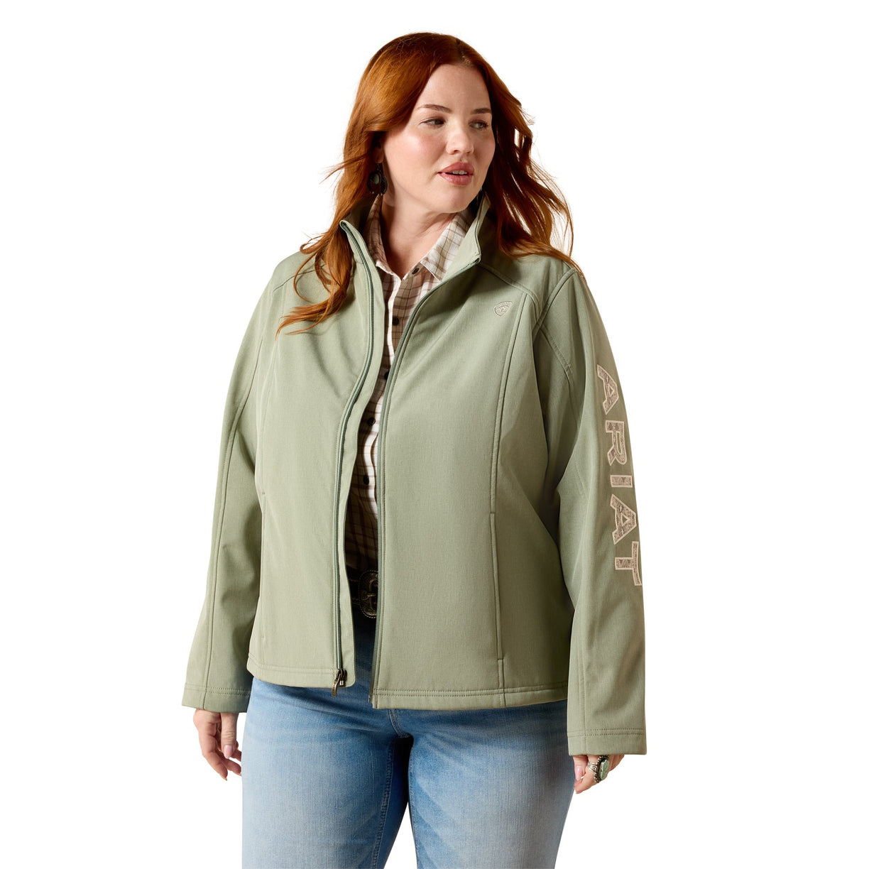 A woman modeling a sage green zip-up jacket with vertical 'AERAT' branding on the sleeve, paired with a patterned shirt and blue jeans against a white background.