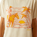 Beige t-shirt with a graphic of a cowboy on a horse, palm trees, and the word 'Ariat' on a beige background.